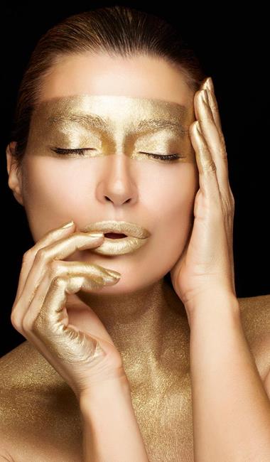 Donna con gli occhi chiusi e trucco dorato su occhi, labbra e mani, su uno sfondo nero.