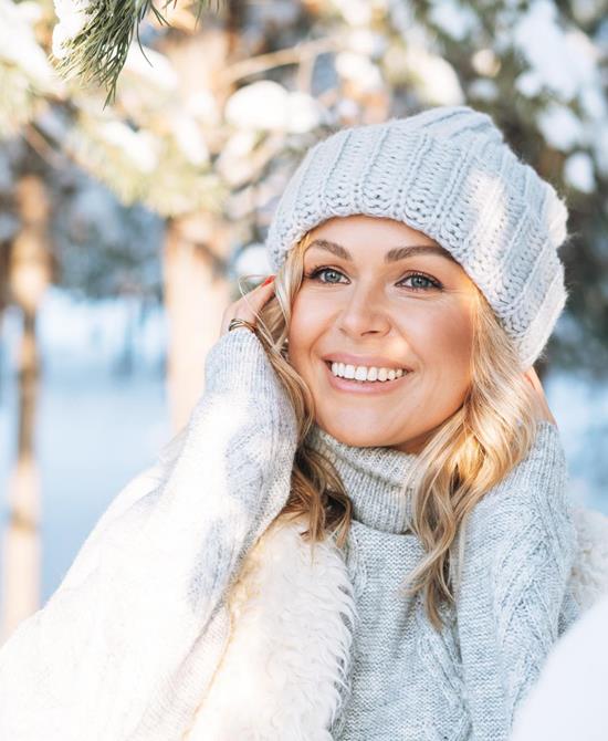 Smiling woman wearing a white knitted hat, scarf and winter clothing in a snow-covered forest.