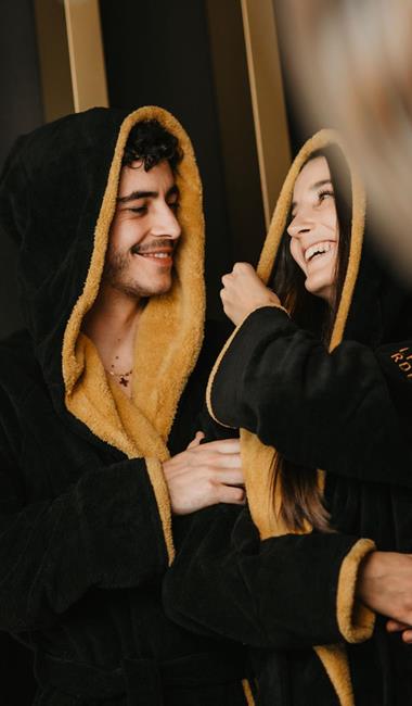 Mann und Frau im schwarz-goldenen Bademantel halten sich lächelnd im Arm.